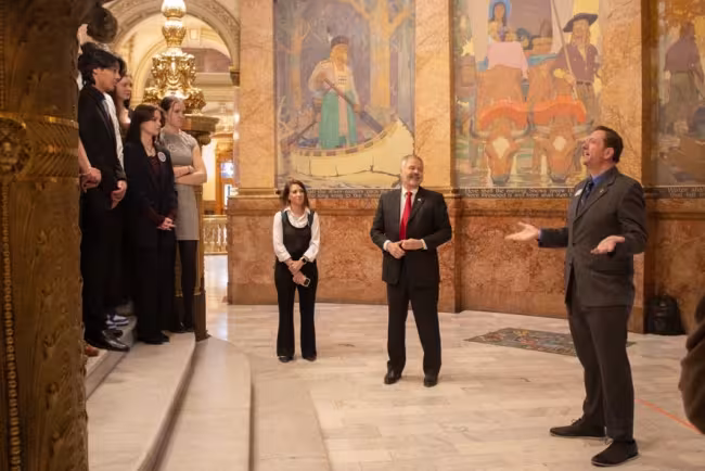 Colorado DMV Driver Services Director Benjamin Mitchell, a man in a grey suit, gestures with his hands while speaking to a group of high school students standing on marble stairs. They are inside the Colorado State Capitol, surrounded by ornate gold pillars and large historical murals. A woman in a black vest stands nearby listening.