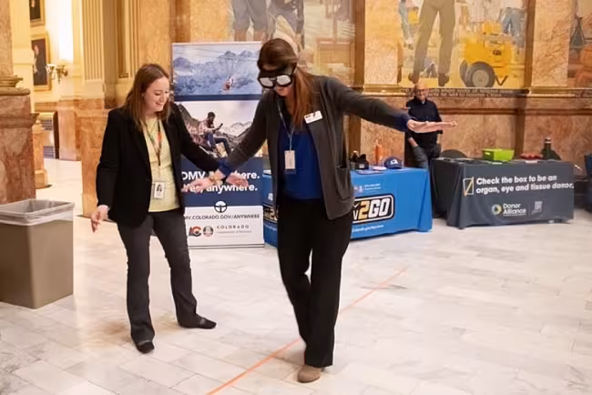 wo female Colorado DMV employees participate in an impairment simulation. One woman wears large black "drunk goggles" and balances with her arms out while attempting to walk a straight orange line on the marble floor. The second woman stands beside her, smiling and offering support. In the background are DMV informational banners and a "Donor Alliance" table.