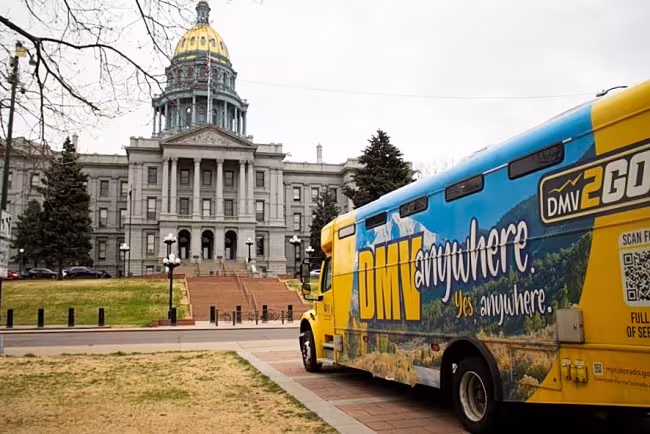 A colorful "DMV2GO" mobile office bus is parked in the foreground, featuring a blue and yellow wrap with the slogan "DMV anywhere. Yes, anywhere." The iconic Colorado State Capitol building, with its distinctive gold dome and classical grey stone architecture, stands prominently in the background under a slightly overcast sky.