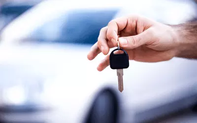 blurry white car in the background and a hand holding a single car key in the foreground