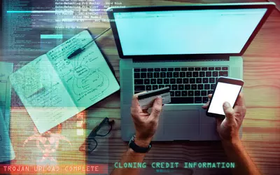 Open laptop with hands holding a smartphone and credit card, there is computer code across the background and the words cloning credit information